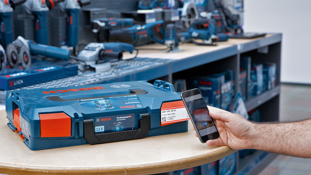 A person scans a blue toolbox on a table with a smartphone. In the background, there are more tools.
