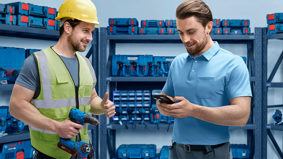 A craftsman wearing a yellow helmet and green vest smiles and gives a thumbs up while holding a drill. Another man in a blue polo shirt looks at his smartphone. In the background, there are shelves with blue toolboxes.