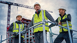 Three construction workers in hard hats and safety vests are smiling at the construction site.
