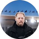 A man with a beard and black jacket stands under floodlights.