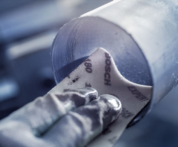 A hand holds Bosch sandpaper against a round metal piece.