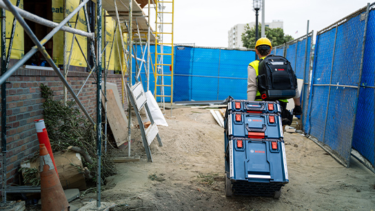 A construction worker wearing a yellow hard hat is pulling a toolbox on a construction path.